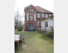 Backsteinbau mit Satteldach und blauen Doppeltüren, daneben ein weißes Holzgebäude. Der Vordergrund zeigt eine Rasenfläche mit Holztisch und klappbaren Stühlen. Im Hintergrund stehen Bäume und vereinzelte Gebäude.