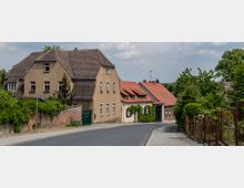 Straße in einem Dorf mit Wohnhäusern auf beiden Seiten, darunter ein großes Gebäude mit beigem Putz und ein kleineres Haus mit roten Dachziegeln. Die Umgebung ist von Bäumen, Büschen und einem Metallzaun begrenzt, bei bewölktem Himmel.