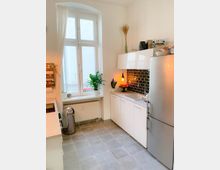 Helle, moderne Küche mit grauem Fliesenboden, weißen Schränken und einer Edelstahlkühlschrank. Ein großes Fenster mit weißen Vorhängen lässt Tageslicht herein, und eine Arbeitsfläche mit Pflanzen, einer Hängelampe und einer schwarzen Fliesenrückwand verleiht dem Raum einen dekorativen Akzent.