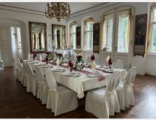 Eleganter Speisesaal mit einer langen, festlich gedeckten Tafel, umgeben von weißen, mit Schleifen dekorierten Stühlen. Der Raum hat verzierte Fenster mit halbtransparenten Vorhängen, goldgerahmte Spiegel und Wandgemälde, einen hölzernen Boden und einen prunkvollen Kronleuchter.