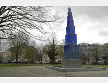Ein öffentlicher Platz mit einem hohen, modernen Denkmal aus blauen, transparenten Elementen auf einem grauen Steinsockel. Im Hintergrund sind Bäume mit kahlen Ästen, Wiesenflächen, Sitzbänke und umliegende Gebäude einer Stadt zu sehen.