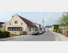 Straßenszene in einer Wohngegend mit überwiegend zweistöckigen Häusern, einem größeren freistehenden Gebäude im Hintergrund und einem Funkmast. Am Straßenrand sind geparkte Autos sowie ein gepflasterter Gehweg und einzelne Bäume zu sehen.