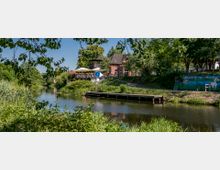 Backsteingebäude mit einem Restaurant und großer Terrasse mit Sonnenschirmen am Ufer eines ruhigen Flusses, umgeben von Bäumen und Grünflächen. Im Vordergrund ein Holzsteg am Wasser und eine bemalte Mauer mit einem Wandbild rechts.