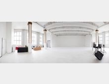 Heller, großzügiger Industriewohnraum mit hohen Decken, weißen Wänden, großen Sprossenfenstern und freiliegenden Stahlträgern. Der Raum enthält eine Sitzecke mit Sofa und Sesseln sowie Fotoausrüstung auf Stativen.