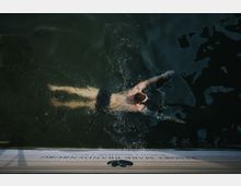 Ein Mann schwimmt im dunklen Wasser neben einem hölzernen Steg, auf dem der Schriftzug „Resort Mark Brandenburg“ zu erkennen ist.