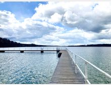 Ein Holzsteg mit weißem Geländer führt auf einen See hinaus, umgeben von bewaldeten Ufern im Hintergrund unter einem Himmel mit Wolken und blauem Himmel.