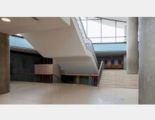Innenansicht eines modernen Gebäudes mit Treppenhaus, das diagonal durch die Mitte des Raums verläuft. Betonpfeiler, große Fensterfronten und Beschilderung zu Bereichen wie Auditorium, Buchladen, Theatersaal und Restaurant prägen den offenen Raum.
