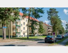 Wohnstraße mit Mehrfamilienhäusern, die gelbe Fassaden und rote Satteldächer haben, sowie Balkone mit grünen Rahmen. Links stehen Kiefern, rechts parken Autos entlang der Straße, und ein Schild weist auf ein Halteverbot hin.