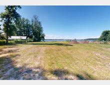 Ein flaches, grasbewachsenes Feld, das auf einen See im Hintergrund blickt, umgeben von Bäumen und einem kleinen Gebäude mit hellen Dächern auf der linken Seite. Rechts befinden sich Erdehaufen und ein Zaun entlang des Feldrandes.