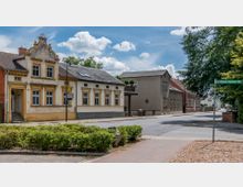 Straßenszene in einem Wohngebiet mit historischen Gebäuden in heller Putz- und Klinkeroptik. Im Vordergrund ist ein Gehweg mit Grünpflanzen zu sehen, im Hintergrund steht ein Straßenschild mit der Aufschrift „Dr.-Wilhelm-Harnisch-Str.“, umgeben von Bäumen und vereinzelten Häusern.