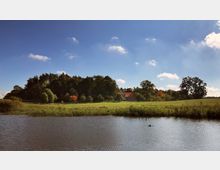 Landschaft mit einem kleinen See im Vordergrund, einer grünen Wiese und Bäumen in der Mitte sowie einem Gebäude mit rotem Dach, das von Bäumen umgeben ist. Der Himmel ist klar und blau mit vereinzelten Wolken.
