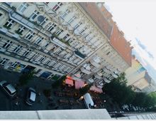 Ansicht einer städtischen Straße aus der Vogelperspektive mit ornaten Altbau-Fassaden. Unten sind geparkte Autos, Fahrräder und ein Straßencafé mit Terrassen unter Markisen zu sehen.