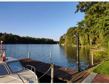 An einem Seeufer befindet sich ein hölzerner Bootssteg mit einem festgemachten Motorboot. Im Hintergrund erstreckt sich ruhiges Wasser, umgeben von dicht bewaldeten Ufern und vereinzelt stehenden Bäumen, die in der Abendsonne leuchten.