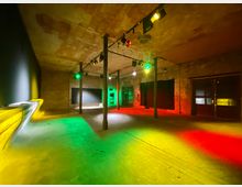 Ein leerer, industrieller Innenraum mit unverputzten Ziegelwänden, einer leicht abgenutzten Beton-/Holzbodenfläche und einer Decke mit sichtbaren Texturen. Der Raum ist mit bunten Stimmungslichtern in Grün, Gelb und Rot beleuchtet, und mehrere Stahlpfeiler und große dunkle Fenster oder Türen sind sichtbar.