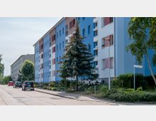 Mehrstöckiges Wohnhaus mit blauer und weißer Fassade sowie farbigen Fensterläden an einer ruhigen Straße. Vor dem Gebäude wachsen Bäume und niedriges Grün, daneben parkende Autos und ein Straßenschild mit der Aufschrift „Am Mühlenweg“.