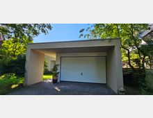 Flacher, moderner Carport mit rechteckiger Form und hellem Putz, integriert in eine grüne Umgebung mit Bäumen und Pflanzen. Die Einfahrt besteht aus einer gepflasterten Fläche, und die Garage hat ein breites, weißes Tor.