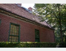 Backsteinfassade eines Gebäudes mit grün umrahmten Fenstern, schrägem Ziegeldach und kleinen Schornsteinen; im Vordergrund eine niedrige Hecke und rechts ein Baum.