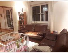 Wohnzimmer mit braunen Ledersofas, einem Bücherregal und großen Fenstern mit Vorhängen. Im Vordergrund steht ein weißes Gitterbett mit buntem Spielzeug, der Boden ist mit Holzparkett ausgelegt.