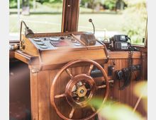 Steuerstand eines Bootes aus Holz mit einem großen Holz-Steuerrad, einem Bedienpult mit analogen Anzeigen und Hebeln sowie Funkgeräten an der Seitenwand. Im Hintergrund sind Fenster mit Blick ins Grüne zu sehen.