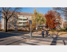 Mehrfamilienhaus aus Ziegelstein mit mehreren Stockwerken und Balkonen, umgeben von Herbstbäumen mit buntem Laub. Im Vordergrund ein Zebrastreifen und zwei Fußgängerinnen, die eine ruhige Straße überqueren.