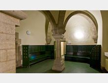 Ein Gewölberaum mit einer zentralen steinernen Säule und Rundbögen, ausgestattet mit dunkelgrünen, an der Wand montierten Sitzbänken. Im Hintergrund führt eine Tür in einen angrenzenden Raum, die Wände sind in hellem Beige gehalten.