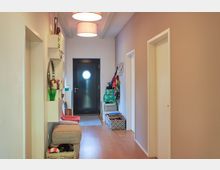 Ein moderner Flur mit Holzfußboden und einer dunkelgrauen Eingangstür mit rundem Fenster. Links befinden sich schmale Regale mit Deko-Objekten und ein runder Spiegel, während rechts Wandhaken für Taschen und eine Ablage mit Körben zu sehen sind.