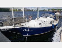 Segelboot mit blauem Rumpf und weißem Deck, benannt "Arielle", liegt an einem Steg in einem kleinen Hafen. Im Hintergrund sind Wasser, Wohngebäude und Bäume sichtbar.