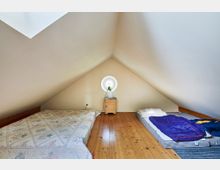 Dachbodenraum mit Holzfußboden, schrägen Wänden und einem runden Fenster an der Stirnseite. Im Raum befinden sich zwei Matratzen mit Bettzeug sowie ein kleiner Holzschrank mit einer Lampe.