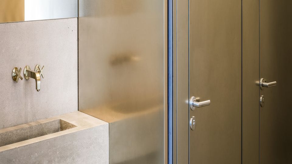 Modernes Badezimmerdetail mit rechteckigem Betonwaschbecken und wandmontierter Armatur neben einer teils matten Glaswand. Rechts sind schmale, metallisch wirkende Türen mit Griffen und Schloss in einem glatten Wandpaneel zu sehen.