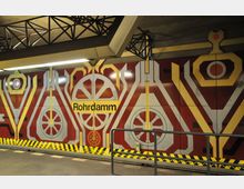U-Bahnhof mit einer bunten Wandgestaltung in roten, gelben und weißen geometrischen Formen; in der Mitte steht der Stationsname „Rohrdamm“. Oben sind Stahlträger der Decke sichtbar, am unteren Rand gelbe und schwarze Warnmarkierungen.