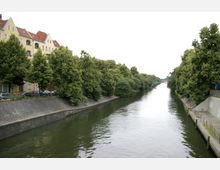 Ein schmaler Fluss mit befestigten Ufern, flankiert von grünen Bäumen auf beiden Seiten; links befindet sich eine Reihe pastellfarbener Wohnhäuser mit roten Dächern, während rechts ein Gehweg mit Geländern verläuft. Im Hintergrund ist eine Brücke zu sehen.
