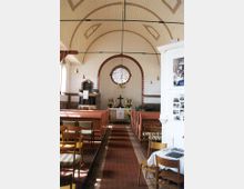Innenraum einer kleinen Kirche mit einem Holzgewölbe. Ein roter Mittelgang führt zum Altar mit einem Kreuz, Blumen und einer Kanzel links daneben. Ein rundes Buntglasfenster und hölzerne Bänke prägen den Raum.