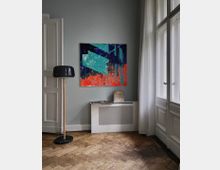 Ein Innenraum mit hellgrauer Wand, darauf ein abstraktes Gemälde in Blau-, Türkis- und Orangetönen. Rechts steht ein großes Fenster mit weißen Vorhängen, links eine offene weiße Tür, unten ein Holzboden im Fischgrätenmuster und ein Heizkörper mit Gitterverkleidung. Ein schwarzer Stehlampenfuß mit Holzstange steht auf der linken Seite.