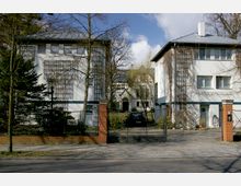 Zwei weiße, zweistöckige Gebäude mit Flachdächern stehen rechts und links eines offenen Torwegs aus Metall und Backsteinpfeilern, dahinter ein Vorgarten mit Bäumen und ein größeres Haus im Hintergrund. Die Fassaden sind mit Klettergittern und Fenstern versehen, und der Bereich ist von vereinzeltem Baumbewuchs umgeben.