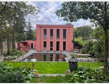 Ein zweistöckiges, rosafarbenes Gebäude mit großen, schwarzen Fenstern steht inmitten eines Gartens. Im Vordergrund befinden sich ein rechteckiger Teich, gepflegte Grünflächen sowie viele Pflanzen, umrahmt von Bäumen und Sträuchern.