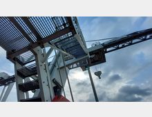 Industriekran mit Metalltreppen und Laufstegen aus Gitterrosten, aufgenommen aus einer Untersicht. Im Hintergrund wolkiger Himmel mit teilweisem Sonnenschein.