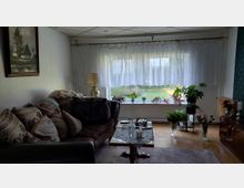 Gemütliches Wohnzimmer mit großem Fenster und weißen Gardinen, davor mehrere Zimmerpflanzen auf der Fensterbank und daneben. Links steht ein gepolstertes Sofa mit Kissen und ein Couchtisch auf Teppich, rechts eine Kommode; der Raum hat Holzfußboden und eine Decke mit Rasterplatten.