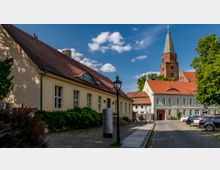 Historischer Dorfplatz mit gepflasterter Straße, flankiert von Gebäuden mit roten Ziegeldächern und hellen Fassaden; im Hintergrund ein Kirchturm aus Backstein mit grüner Spitze. Bäume und Laternen schaffen eine idyllische Atmosphäre.