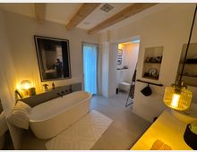 Modernes Badezimmer mit freistehender, weißer Badewanne vor einer beige gestrichenen Wand, die mit einem großen gerahmten Foto dekoriert ist. Der Raum enthält Holzbalken an der Decke, eine Glaspendelleuchte und dekorative Regale; im Hintergrund befinden sich eine Toilette und ein beleuchteter Bereich.