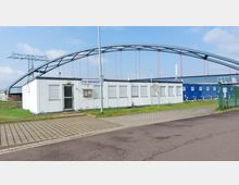 Flaches, weißes Containergebäude mit der Beschriftung „HLB Hafenmeister“ vor einer großen Stahlbogenbrücke. Im Vordergrund sind befestigte Pflasterflächen und im Hintergrund Rasen sowie blau gestrichene Container erkennbar.