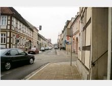 Straßenszene in einer deutschen Altstadt mit zweistöckigen Fachwerkhäusern und Putzbauten auf beiden Straßenseiten. Mehrere parkende und fahrende Autos befinden sich auf der Straße, ein Fußweg ist sichtbar, und Straßenschilder markieren Fahrtrichtungen.