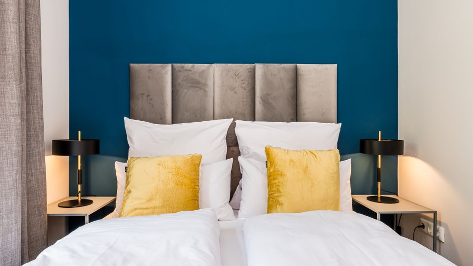 Modernes Schlafzimmer mit zwei nebeneinanderstehenden Betten, weißer Bettwäsche und goldfarbenen Zierkissen vor einem gepolsterten grauen Kopfteil. Dahinter eine dunkelblaue Akzentwand, links und rechts Nachttische mit schwarzen Tischlampen und ein grauer Vorhang am Fenster.