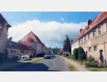 Straße in einem ruhigen Wohnviertel mit Reihenhäusern im traditionellen Stil, gekennzeichnet durch rote Satteldächer und helle Fassaden. Entlang der Straße stehen parkende Autos und gepflegte Vorgärten, im Hintergrund sind Bäume und blauer Himmel mit Wolken sichtbar.