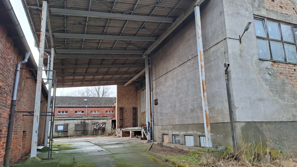 Überdachte Ladezone eines Industriegebäudes mit hohem Metallvordach auf Stahlstützen und Betonboden zwischen Ziegel- und Betonwänden. Im Hintergrund sind weitere niedrige Fabrikgebäude mit Fensterreihen und teils zugemauerten Öffnungen zu sehen.