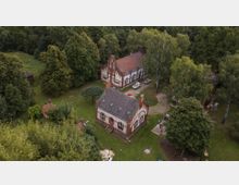 Luftaufnahme eines Anwesens mit historischen Gebäuden im neugotischen Stil, umgeben von Bäumen und Grünflächen. Im Vordergrund ein kleines Ziegelgebäude mit Spitzbögen, im Hintergrund ein größeres Gebäude und parkende Autos auf einem gepflasterten Weg.