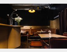 Kleines Café mit modernem Interieur, dunklen Wänden und sichtbaren Lüftungsrohren an der Decke. Es gibt Tische mit braunen Polsterstühlen, dekoriert mit kleinen Vasen und Blumen, sowie eine abgerundete Theke mit einer Vase voller Blumen.