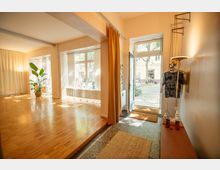 Heller Innenraum mit Holzboden, großen Fenstern auf Straßenniveau, offenem Eingang mit Vorhängen und einer Topfpflanze in der Ecke. Rechts hängt Kleidung neben einem Regal mit verschiedenen Gegenständen, während der Außenbereich durch die offene Tür sichtbar ist.