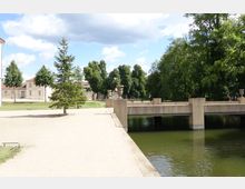 Weitläufiger Park mit einer Brücke aus Beton über einem Wassergraben. Im Hintergrund stehen Bäume und Gebäude mit hellen Fassaden und roten Dächern, daneben eine begrünte Rasenfläche und mehrere Fußwege.