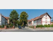 Straßenansicht eines Wohnviertels mit historischen Reihenhäusern, die weiß gestrichen und mit roten Zierelementen versehen sind, umgeben von kleinen Vorgärten und Bäumen; mehrere geparkte Autos entlang der Straße. Ein Zebrastreifen und ein Verkehrsschild sind im Vordergrund sichtbar.