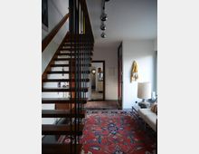 Innenansicht eines modernen Wohnbereichs mit einer offenen Holztreppe und Metallgeländer links. Ein großer, farbiger Orientteppich bedeckt den Boden, rechts stehen eine Couch mit gestreiften Kissen und eine Tischlampe auf einem Beistelltisch. Im Hintergrund führt ein Flur zu einem weiteren Raum.